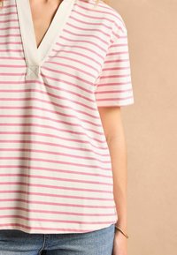 Īsām piedurknēm tops ar V veida kakla izgriezumu, krēmkrāsas fons ar rozā horizontālām svītrām, gluda tekstūra un sānu šķēlumi apakšmalā.