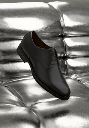 Zwarte leren nette schoen met een gladde afwerking, ronde neus en vijf vetersluitingen, rustend op een metallic zilveren textuur.