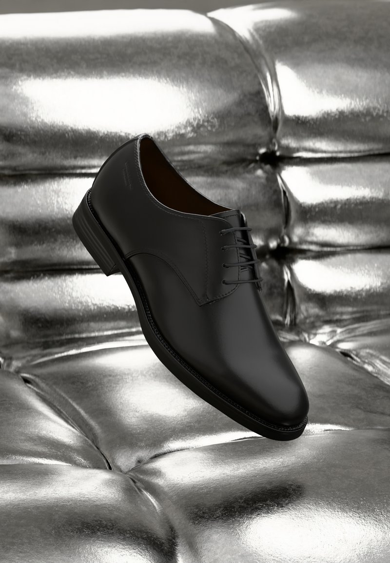 Chaussure de mariage en cuir noir avec une finition lisse, bout arrondi et cinq œillets pour les lacets, reposant sur une surface texturée argent métallique.