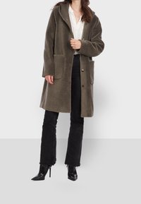 Cappotto in pile verde oliva con cappuccio, due tasche frontali, chiusura a bottone e vestibilit�à comoda. Abbinato a pantaloni neri a zampa d'elefante e tacchi.