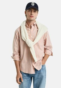 Lichtroze en blauwe gestreepte overhemden met een witte gebreide trui om de nek, gecombineerd met denim jeans en een marineblauwe pet.
