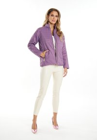Giacca trapuntata lilla con colletto alto, zip frontale e tasche laterali; abbinata a pantaloni color crema e tacchi alti rosa.