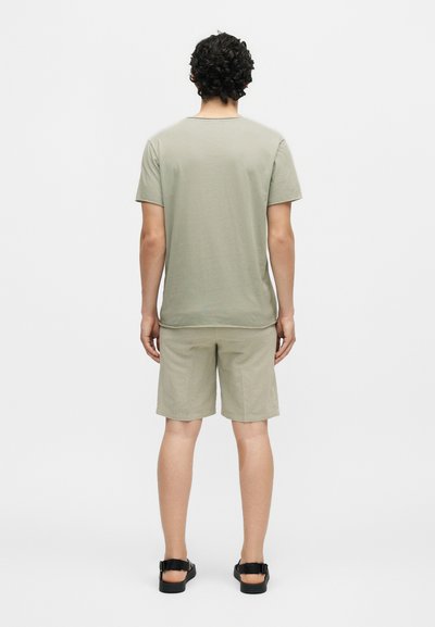 Άνδρας με σγουρά μαλλιά στέκεται με την πλάτη, φοράει ανοιχτοπράσινο t-shirt και ταιριαστό σορτς με μαύρα σανδάλια σε απλό φόντο.