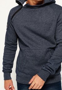 Granatowa bluza z kapturem z miękkiego materiału, z wysokim kołnierzem, zamkiem z przodu, kieszenią kangurka oraz ściągaczami na mankietach. Z czarnym akcentem w postaci przeszyć.