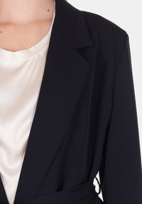 Blazer nero con una texture liscia e strutturata, dotato di un colletto a punta e una cintura alla vita, indossato sopra una blusa crema.