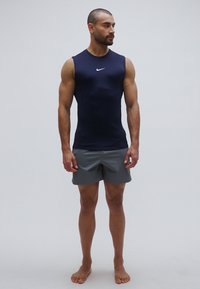 Top sportivo senza maniche blu navy con logo bianco, abbinato a pantaloncini grigio chiaro. Il tessuto appare liscio e aderente, ideale per attività sportive.