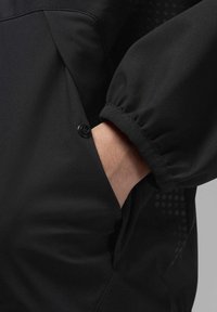 Chaqueta negra con un bolsillo frontal con cierre de presión, puños de manga elásticos y acentos texturizados en los lados. Acabado satinado.
