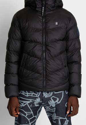 Mand iført en sort polstret jakke med lynlås og lommer foran, kombineret med mørkeblå jeans med et lyst abstrakt mønster.