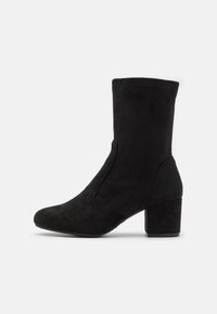Bottine en daim noir avec un talon épais, bout pointu et panneaux latéraux en élastique. Texture lisse sans motifs ni accents visibles.