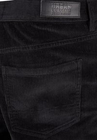 Gros plan sur un pantalon en velours côtelé noir montrant la poche arrière et l’étiquette en cuir avec la marque Urban Classics Denim Factory.