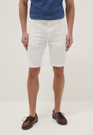 Man draagt witte knielange shorts, een blauw overhemd en donkerbruine veterschoenen, staand tegen een effen lichte achtergrond.