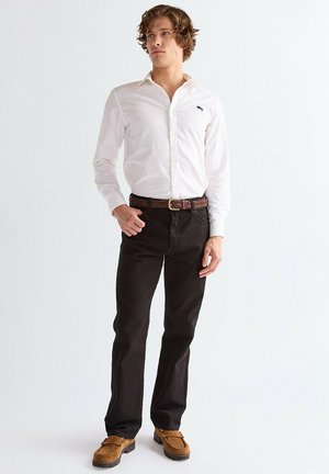 Hombre con camisa blanca de manga larga abotonada, pantalones oscuros, cinturón trenzado marrón y zapatos de ante marrones, de pie contra un fondo blanco.