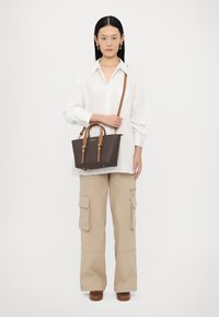 Bruine handtas met een patroon, voorzien van lichtbruine handvatten en een schouderriem, gecombineerd met een witte blouse met knopen en beige cargo-broek. Effen achtergrond.