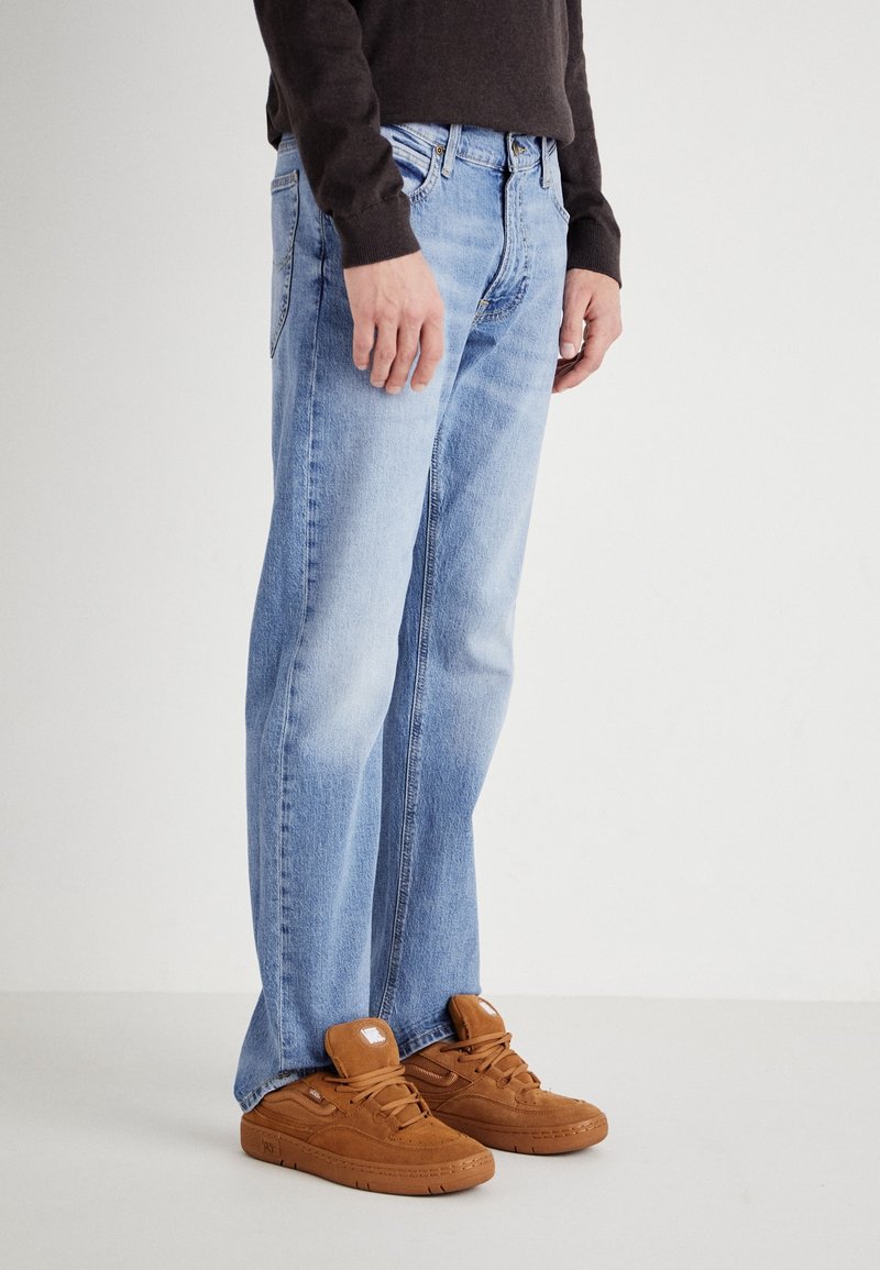 Personne portant un jean bleu clair, des baskets marron et un haut à manches longues foncé, debout sur un fond uni.