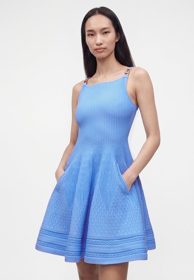 Femme aux longs cheveux foncés portant une robe bleue texturée sans manches avec des poches et des bretelles décoratives sur fond blanc.