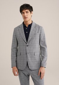 Grijze geruite blazer met opstaande revers, twee voorzakken en een enkele knoopsluiting, gedragen over een marineblauwe poloshirt.