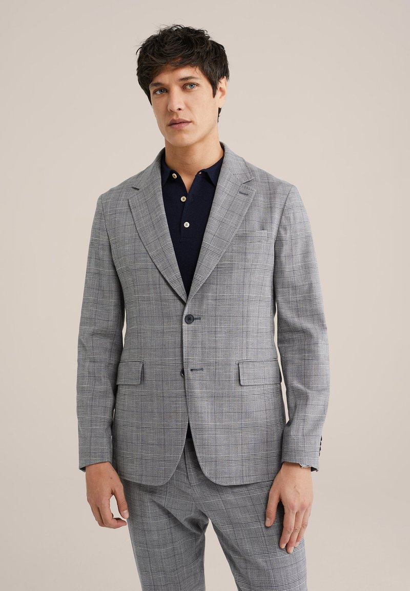 Grijze geruite blazer met opstaande revers, twee voorzakken en een enkele knoopsluiting, gedragen over een marineblauwe poloshirt.