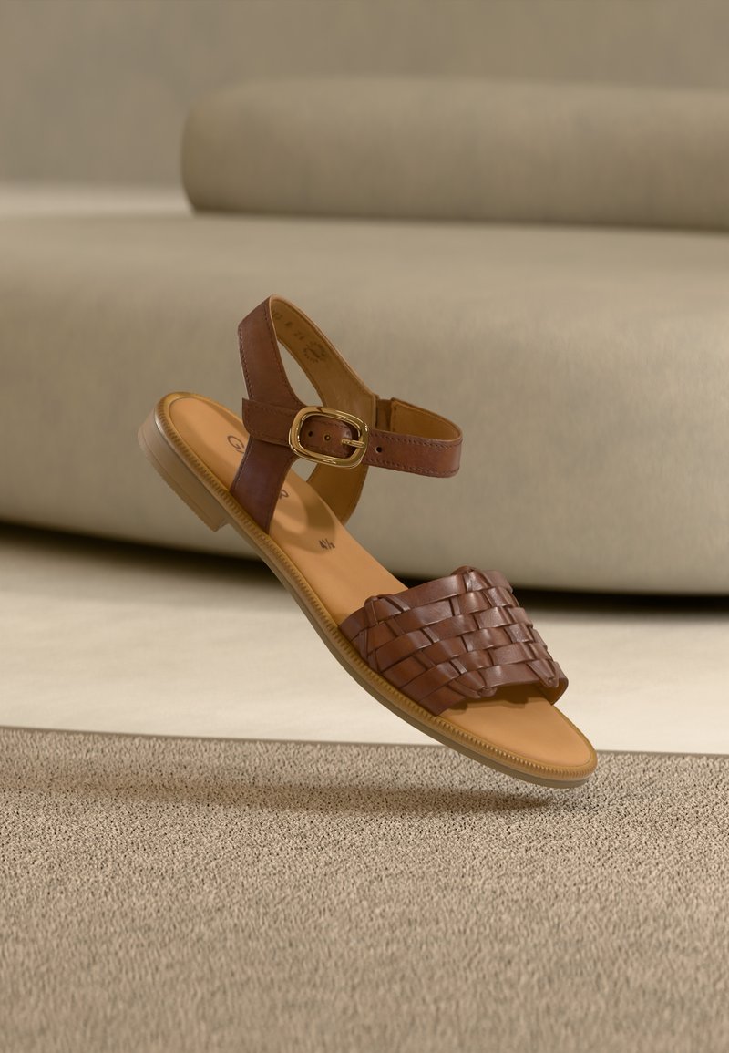 Καφέ δερμάτινο σανδάλι με πλεγμένο σχέδιο λουριού, λουρί αστραγάλου και στρογγυλεμένη μύτη. Διαθέτει επίπεδη σόλα και χρυσή αγκράφα.