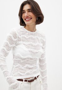 Top bianco a maniche lunghe realizzato in pizzo trasparente, con una texture ondulata e dettagli sfrangiati. Abbinato a pantaloni chiari e una cintura marrone.