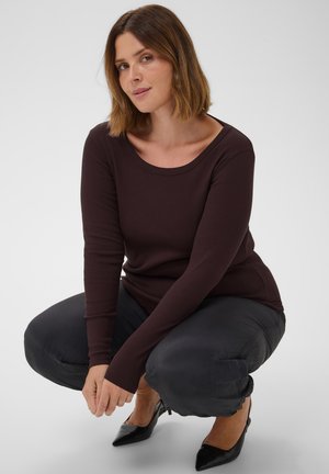 Femme aux cheveux mi-longs accroupie, vêtue d'un haut à manches longues marron foncé, d'un pantalon noir et de talons pointus noirs sur fond blanc.