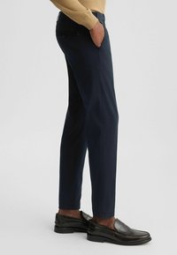 Navy Hosen im geraden Schnitt, aus glattem Stoff, mit seitlichen Taschen und einer geknöpften Gesäßtasche, kombiniert mit schwarzen Slippern.