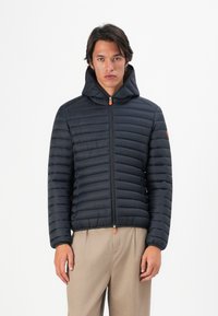 Jonge man in een zwart gewatteerd jack met capuchon en rits, en beige broek, staande tegen een witte achtergrond.