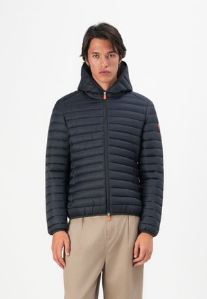 Young man wearing a black quilted hooded jacket with zipper and beige pants, standing against a white background.