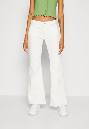 Osoba nosząca białe rozszerzane dżinsy, białe trampki i zielony krótki, bezrękawnikowy top, stojąca na jednolitym tle.