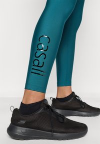 Τιρκουάζ κολάν με λαμπερό μαύρο λογότυπο στο πλάι, συνδυασμένα με μαύρα sneakers από δίχτυ  με σόλα αφρού και κορδόνια.