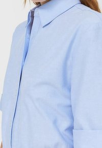 Camicia a maniche lunghe di colore blu chiaro realizzata in tessuto morbido, con colletto classico e maniche risvolte, dal taglio aderente e con una leggera texture.