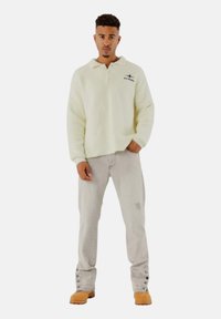 Flauschiger, cremefarbener Pullover mit Kragen und Logo, kombiniert mit hellgrauen Jeans mit seitlichen Knöpfen. Braune Stiefel vervollständigen das Outfit.