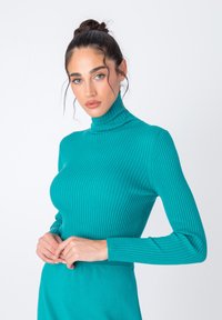 Turkizna rebrasta pulover s turtleneck ovratnikom in dolgimi rokavi, prilega se in je izdelan iz teksturirane tkanine. Oblečen z rokami prepletenimi okoli pasu.