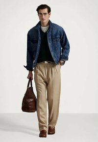 Mężczyzna w okularach, w dżinsowej kurtce, ciemnym swetrze, beżowych spodniach i brązowych butach, trzymający dużą brązową skórzaną torbę w lewej ręce.