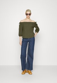 Top verde-oliva sem alças com um acabamento em babados e mangas amarradas. Combinado com jeans de perna larga azul-escuro e sapatos de salto plataforma em tom caramelo.