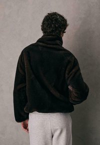 Mørkebrune faux fur jakke med høj krave og lange ærmer, der har en blød tekstur. Stylings med grå sweatpants. Bagvejr.