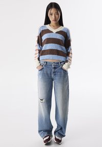 Maglione a strisce in marrone, blu e crema; jeans in denim oversize con strappi; calzature sportive con un accento color bordeaux.