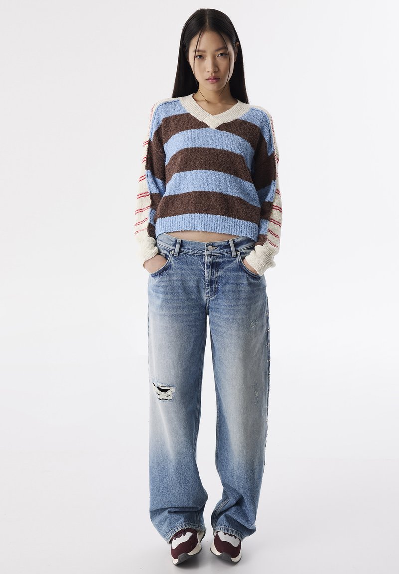 Maglione a strisce in marrone, blu e crema; jeans in denim oversize con strappi; calzature sportive con un accento color bordeaux.