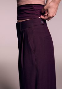 Pantaloni viola a vita alta con pieghe, tasche laterali e chiusura a bottone. Tessuto morbido e liscio. Dettagli inseriti visibili in vita.