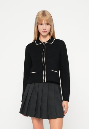 Cardigan noir avec bordure et boutons blancs, doté d'un col et de deux poches avant, associé à une jupe grise plissée.