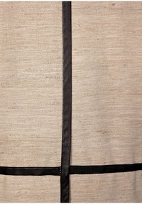 Beige texturerad tyg med svarta läderdetaljer som bildar ett korsmönster. Ytan ser vävd ut med synliga fibrer och små variationer.