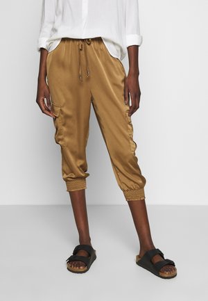 Persona vistiendo pantalones jogger satinados cortos de color camel con puños elásticos y una camisa blanca de manga larga remangada, combinados con sandalias negras.
