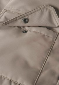 Beige stof med en glat tekstur, der har en trykknaplukning og kontrasterende syninger, fremviser et foldet design med vinklede kanter.