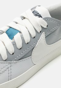 Šedé kožené tenisky s bielymi šnúrkami, so modrým akcentom a bielym logom Nike. Štruktúrovaný zvršok s viditeľnými detailmi šitia.