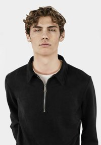 Giacca in suede nera con colletto e zip frontale, caratterizzata da un design semplice e da una texture morbida. Indossata sopra una camicia chiara.