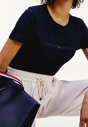 Person iført en marineblå Tommy Hilfiger T-shirt og lyse beige bukser med snøre, der holder en marineblå taske med rød, hvid og blå rem.