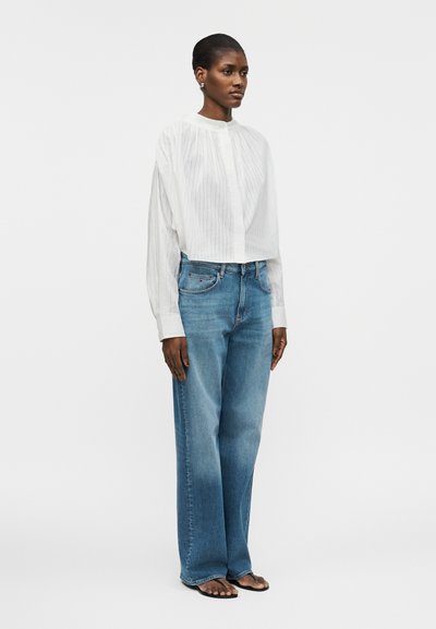 Γυναίκα όρθια που φοράει μια λευκή ριγέ μπλούζα με μακριά μανίκια, τζιν παντελόνι ψηλόμεσο, φαρδύ, σε μπλε χρώμα και μαύρες σαγιονάρες, μπροστά σε λευκό φόντο.