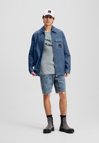 Veste bleue avec une fermeture éclair et une poche poitrine, t-shirt gris avec du texte noir, shorts en denim déchirés, et bottes grises avec des accents noirs.