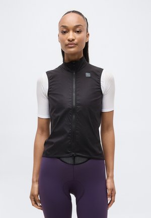 Kvinde stående iført en sort ærmeløs vest med lynlås over en hvid kortærmet top og lilla polstrede cykelshorts på en ensartet baggrund.