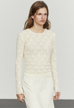 Kvinde iført en hvid langærmet blonder-top og hvid nederdel, der står mod en ensartet lys baggrund.