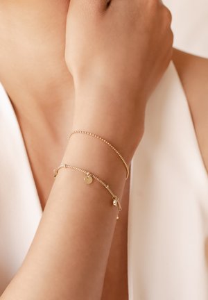 Goldfarbene Armbänder, eines mit kleinen Perlen, das andere mit einem glatten Scheiben-Anhänger und zarter Kette, vor einem hellen Hautton.
